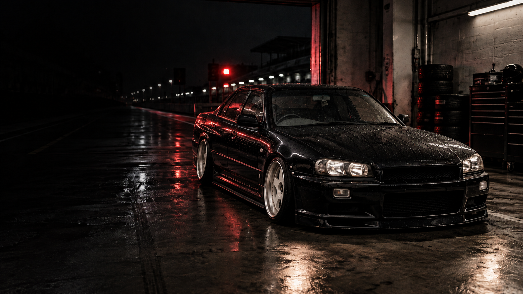 Black R34 sedan under the lights at a rain-soaked pit garage at night